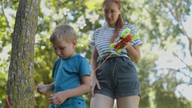 Genç anne, mutlu oğluna parktaki bir ağaç gövdesine halatla nasıl düğüm atılacağını öğretiyor.