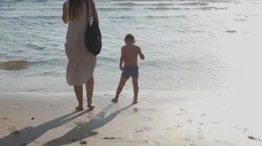 Altın saatinde deniz kıyısında yalınayak yürüyen genç bir anne ve oğlunun arka görüntüsü.