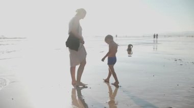 Bir kadın ve küçük bir çocuk deniz kenarında yalınayak yürüyor. Yaz akşamının tadını çıkarıyorlar.