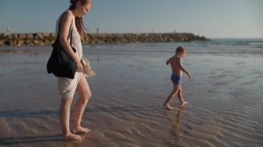 Annesi ıslak kumsalda yanında yürürken genç bir çocuk oynuyor ve suya atlıyor.