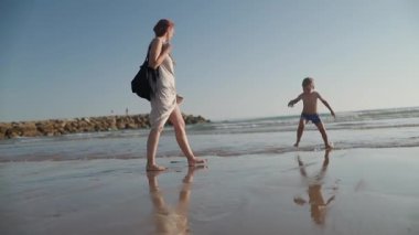 Genç anne, yaz tatilinde oğlunun güneşli bir plajda dalgalar ve kumlarla oynamasını izliyor.