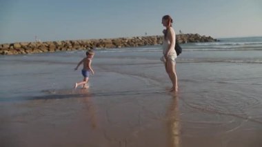 Genç çocuk gün batımında annesiyle kıyı şeridi boyunca koşarken neşeyle su sıçratıyor.