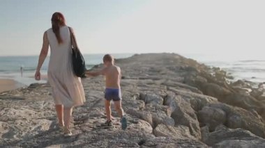Genç kadın ve çocuğu el ele tutuşurken taştan bir dalganın üzerinde denize doğru yürüyorlar.
