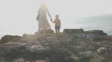 Güneşli bir akşamda okyanus kıyısındaki kayalarda yürürken oğullarının elini tutan genç anne.