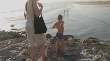 Genç bir kadın ve çocuğu kayalık bir sahil boyunca yürüyor ve birlikte okyanus dalgalarını izliyorlar.