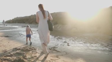 Genç anne ve küçük oğlu güzel bir gün batımında kumlu sahilde yürüyor ve oynuyorlar.