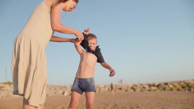 Genç anne çocuğuna bakıyor, gün batımında kumlu bir plajda gömlek giymesine yardım ediyor.