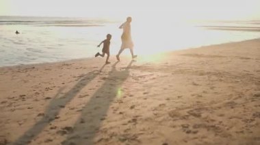 Güzel altın bir günbatımında annesine sarılmak için deniz kıyısında koşan mutlu küçük çocuk.