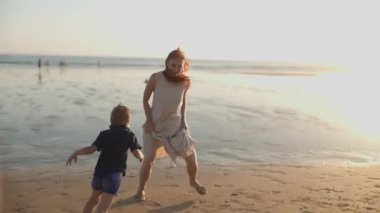 Genç anne güzel bir gün batımında kumlu deniz kıyısında mutlu oğlunu kovalıyor.