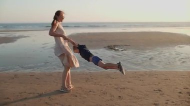 Genç anne güzel altın bir günbatımında kumlu bir sahilde mutlu çocuğunu döndürüyor.