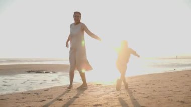 Neşeli anne ve küçük oğlu el ele tutuşup deniz kenarındaki kumda birlikte oynuyorlar.