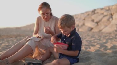 Genç bir çocuk ve annesi kumsalda oturmuş meyve yiyor ve sahilde birlikte oynuyorlar.