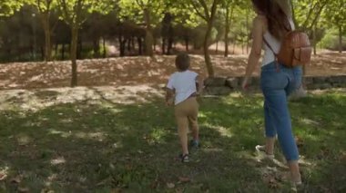 Genç bir kadın ve çocuğu güneşli bir ormanda kameradan uzaklaşıyorlar.