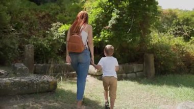 Bir kadın ve küçük bir çocuk, güneşli bir parkta, taş bir yolda el ele yürüyorlar.