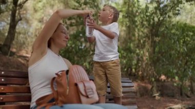 Genç çocuk, annesi birlikte bir bankta oturup izlerken şişeden su içiyor.