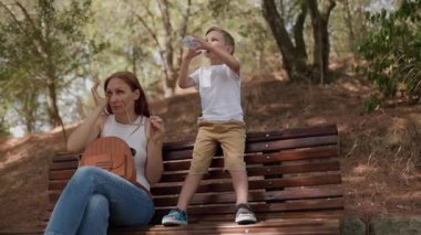 Genç bir aile, güneşli bir günde bankta otururken plastik bir şişeden su içiyor.