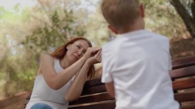 Neşeli anne güneşli bir parkta bankta otururken oğluyla oynuyor ve onunla konuşuyor.