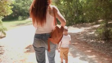 Genç bir anne ve küçük oğlu yazın güneşli bir parkta kirli bir yolda yürüyorlar.