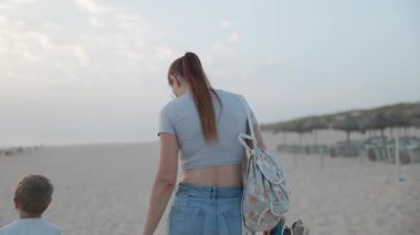 Genç bir kadın ve küçük oğlu el ele tutuşuyor ve alacakaranlıkta kumlu bir sahil şeridinde birlikte yürüyorlar.