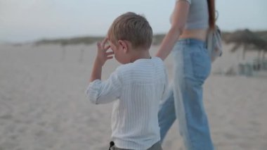 Kumsalda yürürken annesinin elini tutan ve bir şeyi işaret eden genç bir çocuk.