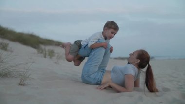 Mutlu anne kumlara uzanıp oğlunu ayaklarıyla kaldırıyor ve eğlenceli bir uçak oyunu oynuyor.