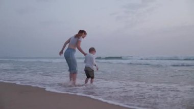 Mutlu bir aile, bir anne ve küçük oğlu, el ele tutuşup, alacakaranlıkta okyanusta dalgalara atlıyorlar.