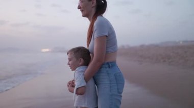 Genç oğluyla birlikte sakin bir akşamın tadını çıkaran bir kadın kumlu bir sahilde hafif dalgaları seyrediyor.