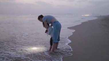 Genç bir anne ve oğlu alacakaranlıkta kumlu bir plajda sığ suyu fenerle aydınlatıyorlar.