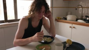 Mutfak masasında tek başına yemek yiyen üzgün ve düşünceli, uzun saçlı bir yetişkin.