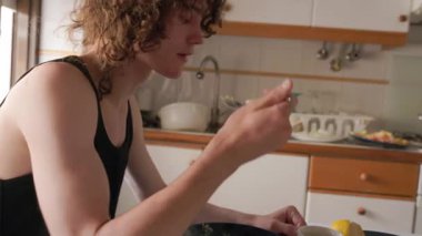 Genç beyaz, kıvırcık saçlı bir adam evcil bir mutfakta kaşıkla sağlıklı bir yemek yiyor.