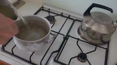 Birisi alüminyum bir tencereye beyaz bir gaz sobasıyla çorba döküp kaşıkla karıştırıyor.