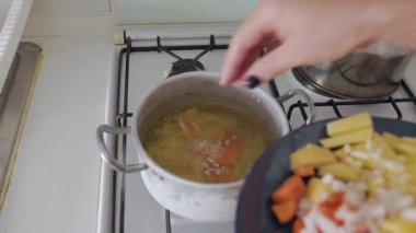 Gaz sobasındaki kaynayan et suyuna taze sebze ekleyen ve karıştıran bir kadın.