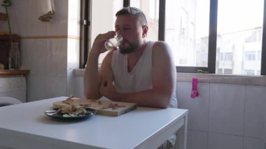 Sakallı bir adam kızarmış ançüez yiyor ve açık bir mutfakta temiz alkol içiyor.