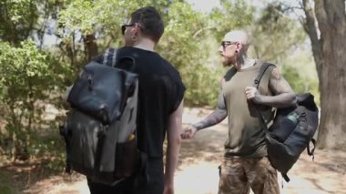 Yeşillik bir ormanda gezen dövmeli ve sırt çantalı iki genç adam.