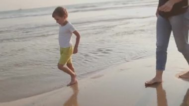 Çıplak ayaklı çocuk annesiyle ıslak kumda yürüyor ve sahilden deniz kabuğu topluyor.