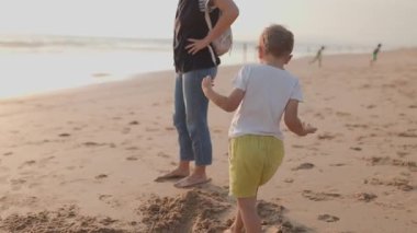 Genç çocuk gün batımında sahilde annesiyle yalınayak yürümeden önce kumda oynuyor.