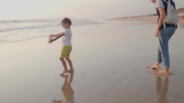 Mutlu anne ve küçük oğlu gün batımında sahilde dalgalarla oynayıp yürüyüş yapıyorlar.