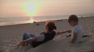 Mutlu anne ve küçük oğlu güzel bir günbatımında kumlu bir kumulda birlikte oynuyorlar.