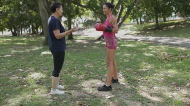 Boks eldivenli genç bir kadın güneşli bir parkta kişisel koçuyla antrenman yapıyor.