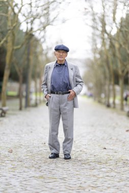 Kendinden emin yaşlı bir adam ağaçlarla kaplı bir kaldırım yolunda duruyor, elleri ceplerinde poz veriyor.