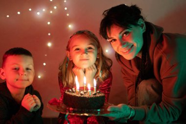 Anne ve çocuklar bir doğum günü kutluyor, ellerinde mumlarla bir pasta tutuyorlar, neşe ve mutluluk dolu.