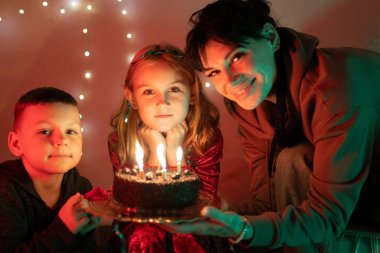Anne ve çocuklar doğum gününü kutluyorlar, ellerinde üç mumla çikolatalı pasta tutuyorlar.