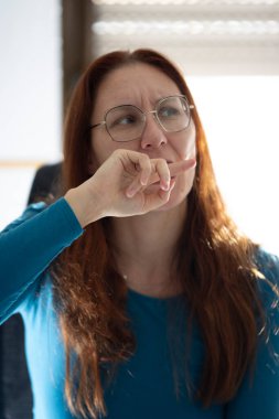 Konsantre olan, gözlüklü ve mavi gömlekli bir kadın, düşünceli bir ifadeyle başka yere bakıyor.