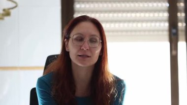 Kızıl saçlı ve gözlüklü genç bir kadın sanal bir konferans sırasında el kol hareketi yapıyor ve konuşuyor.