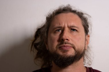 Sakallı ve kıvırcık saçlı bir adam beyaz arka planda sorgulayıcı bir ifadeyle düşünüyor.