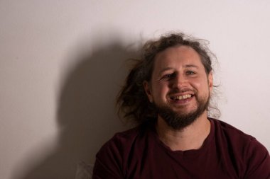 Sakallı ve kıvırcık saçlı neşeli bir adam beyaz bir duvara gülüyor.