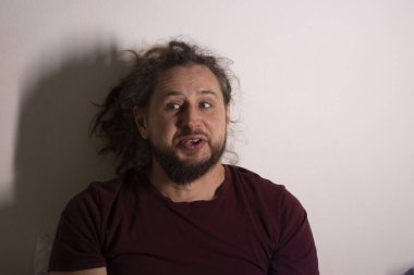 Kıvırcık saçlı ve sakallı bir adam açık arkaplanda şaşırtıcı bir ifade sergiliyor.