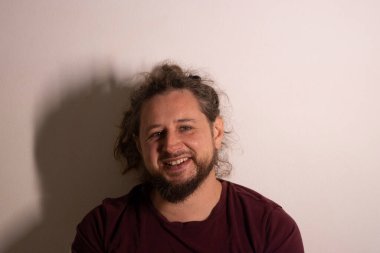 Sakallı bir adam sade bir duvara gülümsüyor, mutluluk ve olumlu duygular yayıyor.