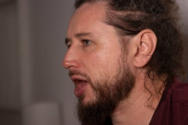 Sakallı ve uzun saçlı bir adam ağzı açık konuşuyor.