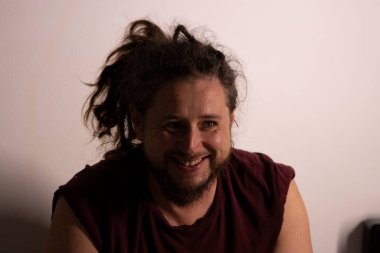 Saçlı ve sakallı gülümseyen adam sade bir arka planda neşeyle gülüyor.